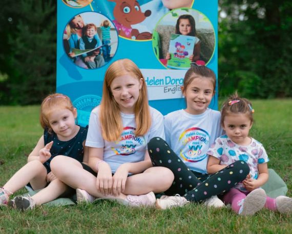 Four smiling children sitting on grass in front of a colorful banner, enjoying a sunny day outdoors.